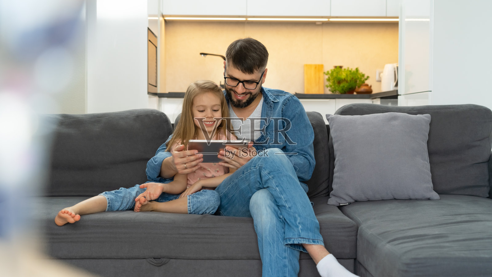 父亲和孩子坐在家里的沙发上使用平板电脑。电脑游戏,上网,社交媒体,在网上商店购买照片摄影图片
