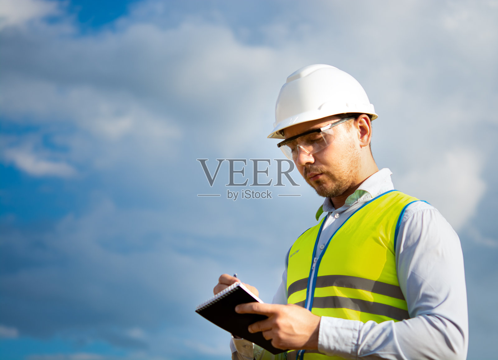 工程师戴着头盔,戴着眼镜,穿着黄色马甲,手上拿着头盔,手里拿着笔记本、文件夹,在笔记本上做笔记照片摄影图片