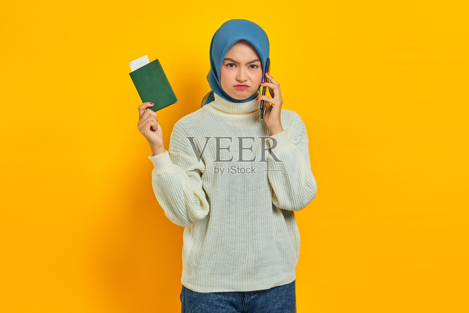 肖像沉思美丽的亚洲女人在白色毛衣通话的手机和持有护照隔离在黄色的背景。夏天的旅行照片摄影图片