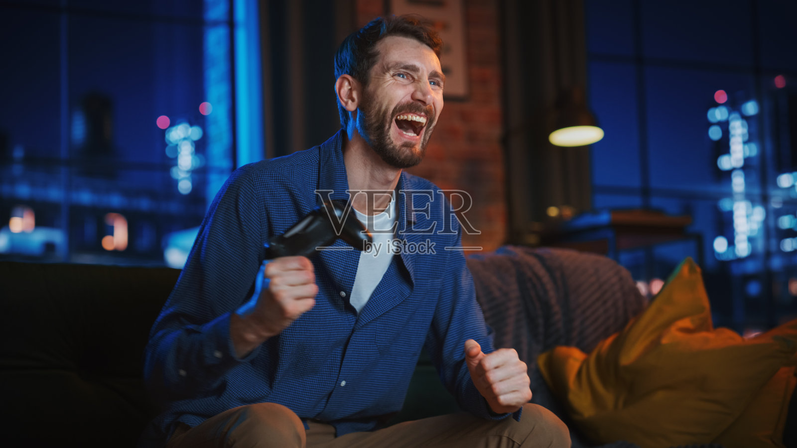 年轻英俊男子的肖像花时间在家里,坐在沙发上在时尚的阁楼公寓和在游戏机上玩视频游戏。成功的男性使用控制器玩动作游戏并获胜。照片摄影图片