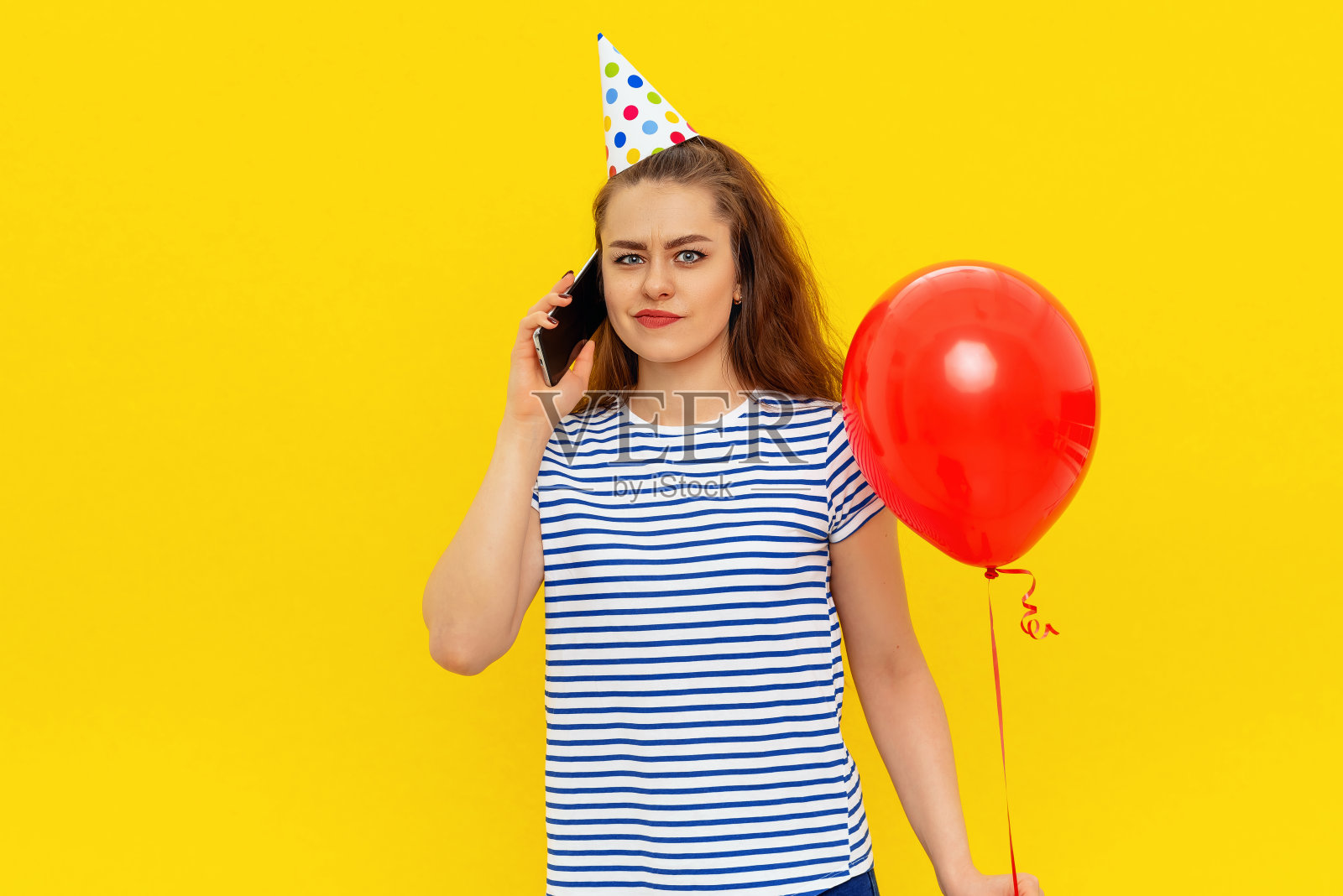 不开心困惑的年轻女子正在讲手机,穿着派对锥形和条纹t恤,感到尴尬和冒犯有不好的心情在庆祝期间,站在黄色的背景照片摄影图片