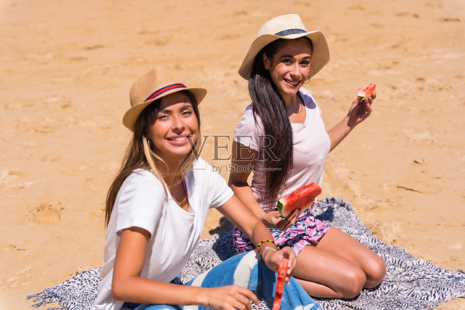夏天的沙滩上,两个朋友坐在沙滩上笑着吃着西瓜,背景是大海照片摄影图片