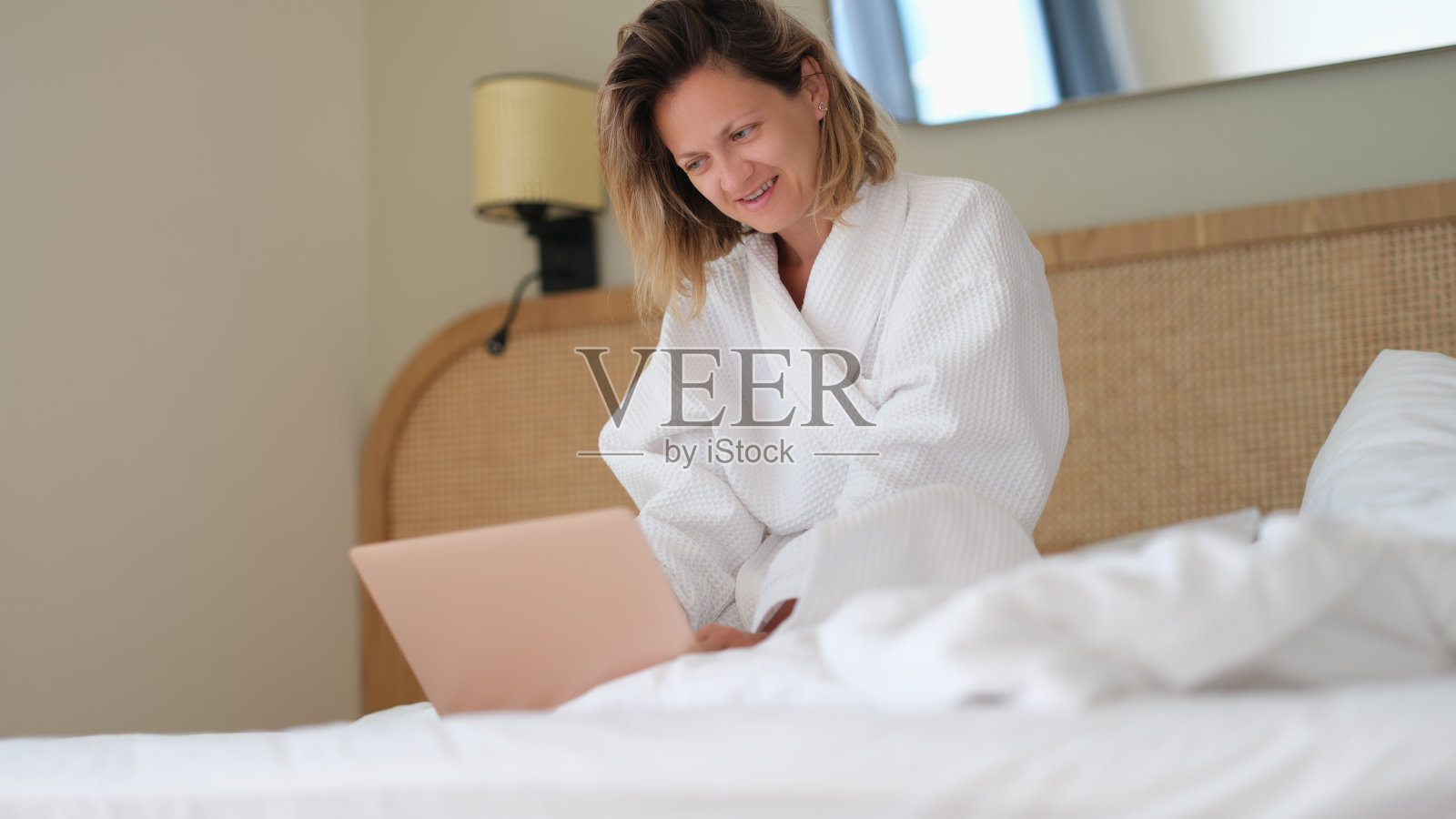 女人坐在家里的床上,穿着白色浴衣,在笔记本电脑上工作照片摄影图片