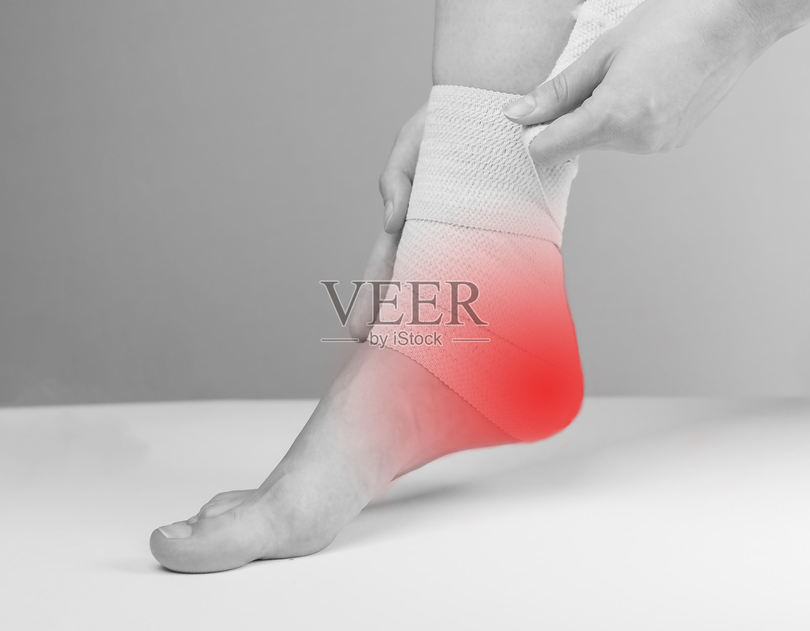 妇女用弹性绷带包裹疼痛的脚踝,以减轻脚后跟疼痛和炎症。脚上有红斑。足底筋膜炎、肌腱炎治疗。卫生保健的概念。黑色和白色照片摄影图片