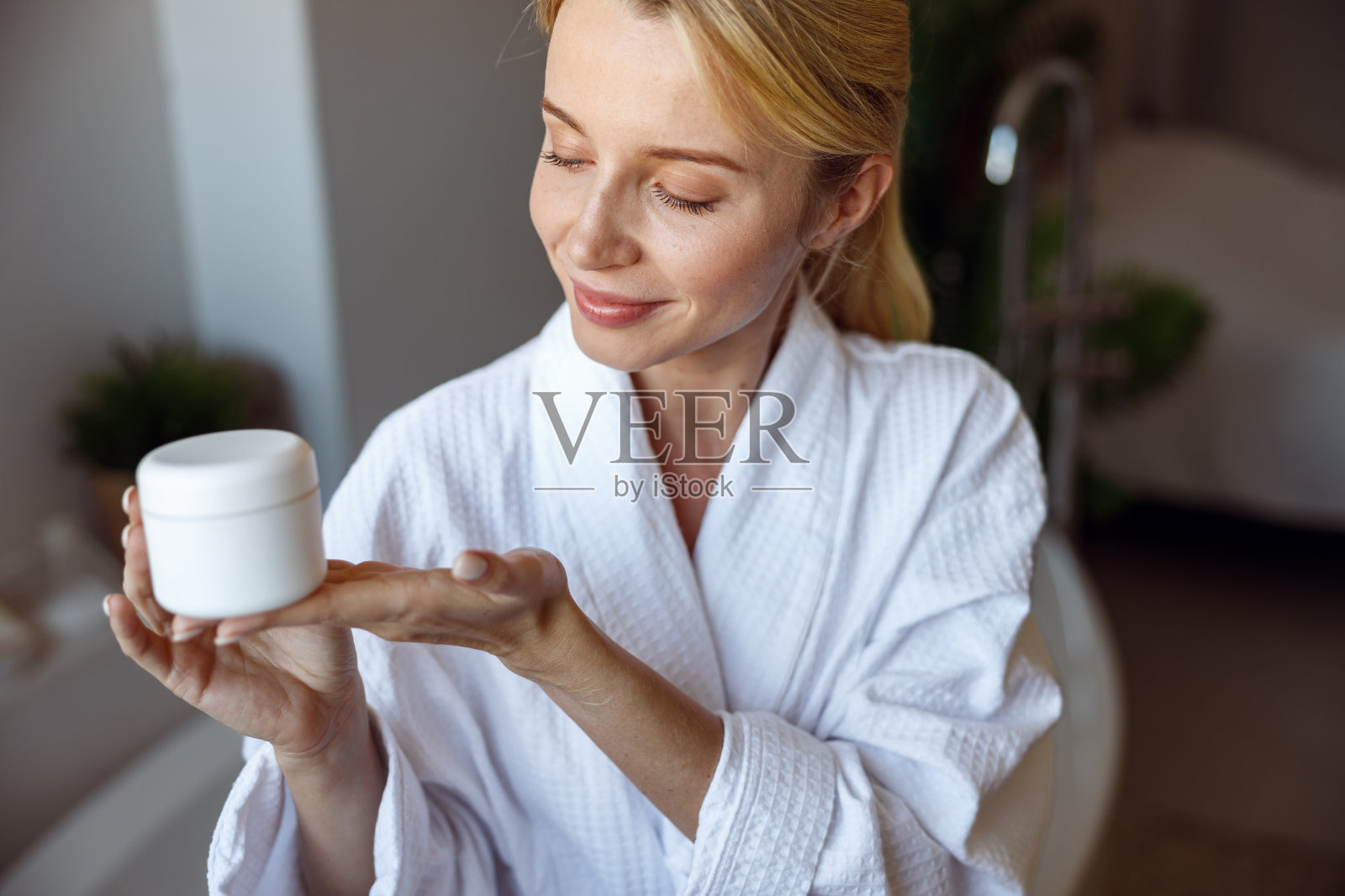 迷人的金发年轻女性展示面霜或身体乳液。护肤习惯。照片摄影图片