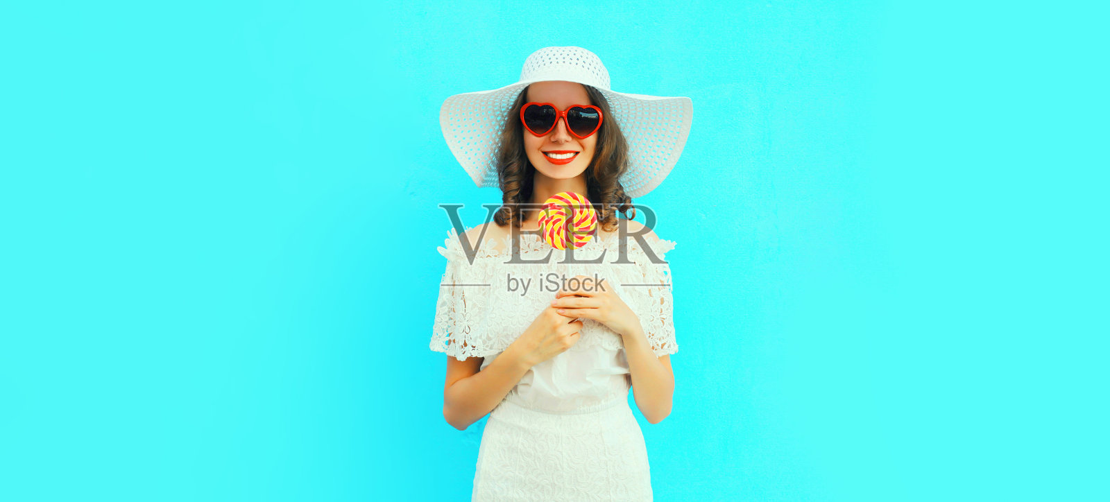 幸福微笑的年轻女子的肖像甜棒棒糖戴着红心形状的太阳镜戴夏天白色的草帽在蓝色的背景照片摄影图片