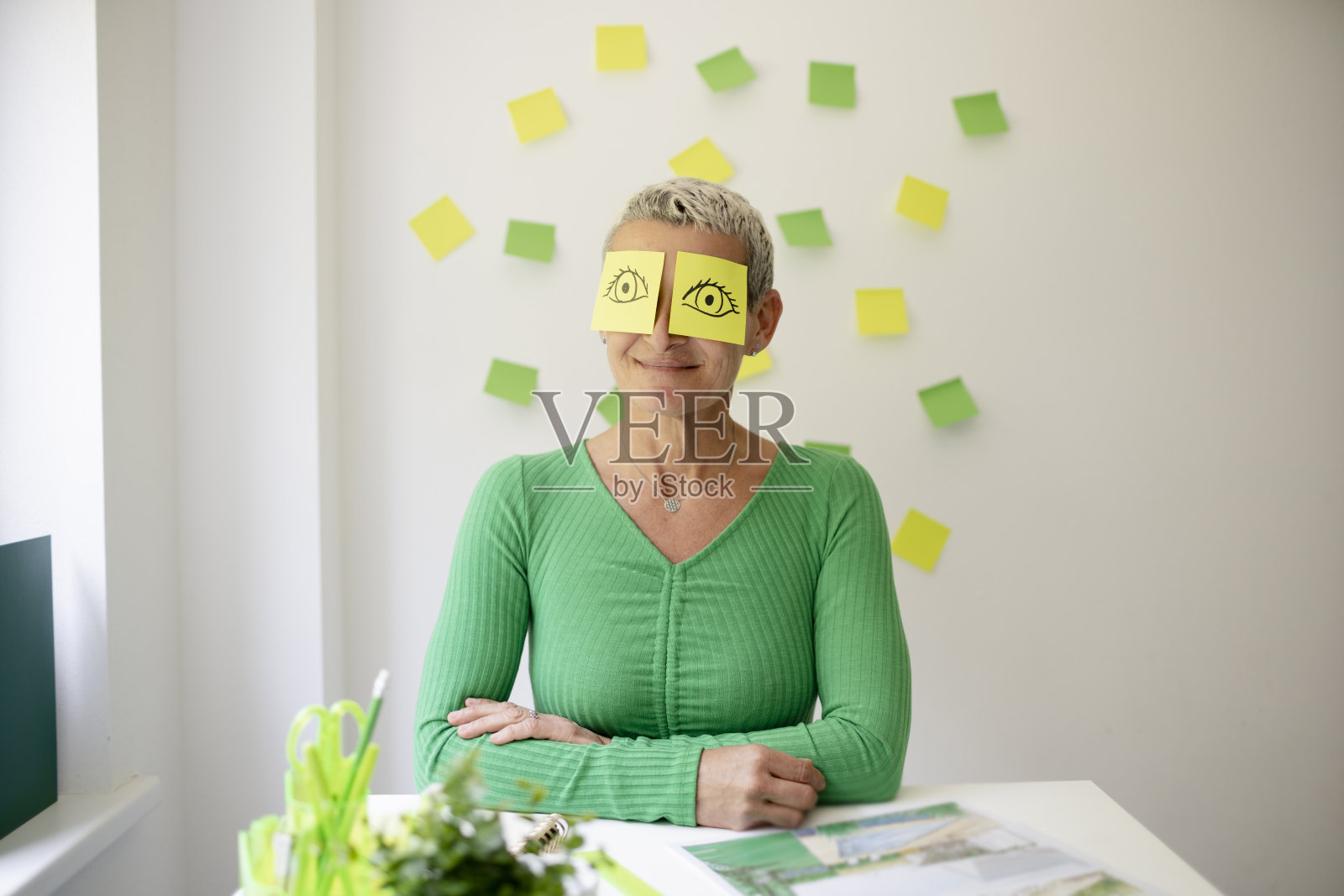 短发的中年妇女坐在摆满绿色植物和工作笔记的办公桌前,脸上画着眼睛的便签纸照片摄影图片