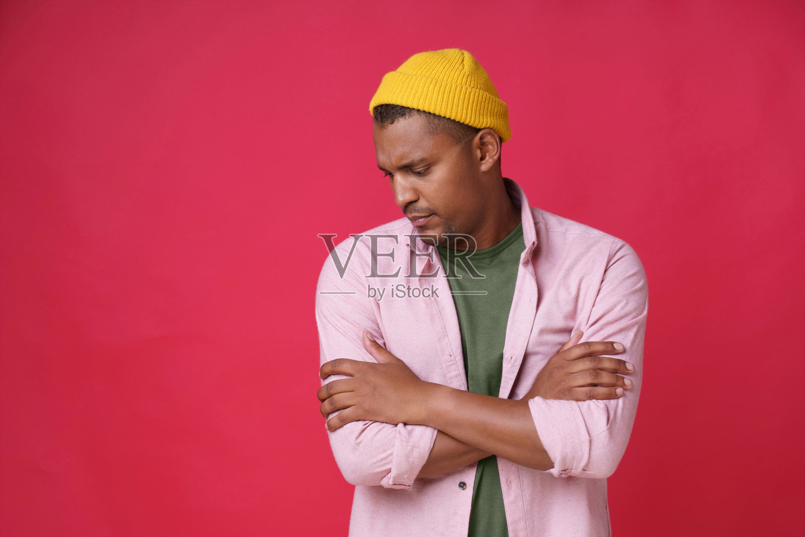严肃热情英俊的非洲裔美国年轻人摆姿势,双手合十,头斜向下,穿着休闲的粉红色衬衫和黄色帽子微笑的相机孤立的红色背景照片摄影图片