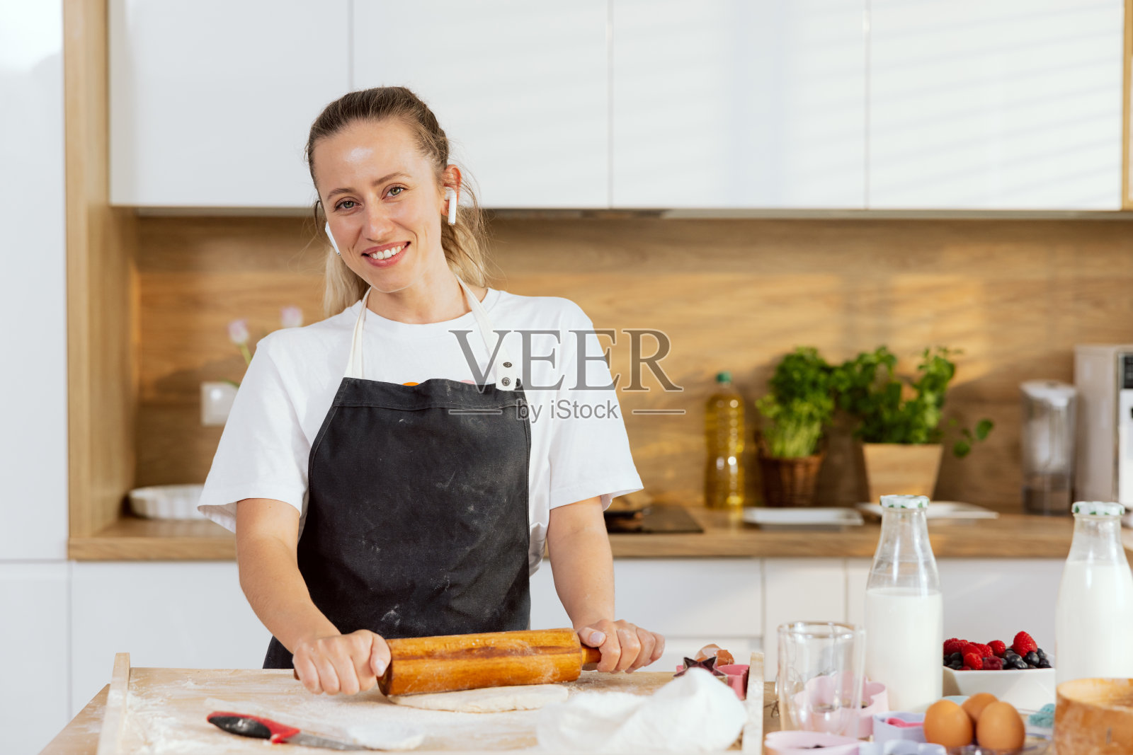 迷人的金发女子站在厨房的桌子上看着镜头,烘烤,烹饪,为面包,披萨,馅饼,饼干准备面团,戴着耳机听音乐照片摄影图片
