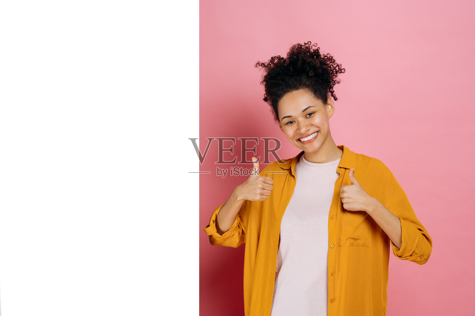 实物模型。兴奋的卷发非洲裔美国女孩,穿着休闲服装,竖起大拇指,站在空白的地方做广告或展示,在粉红色的背景上,看着镜头,微笑照片摄影图片