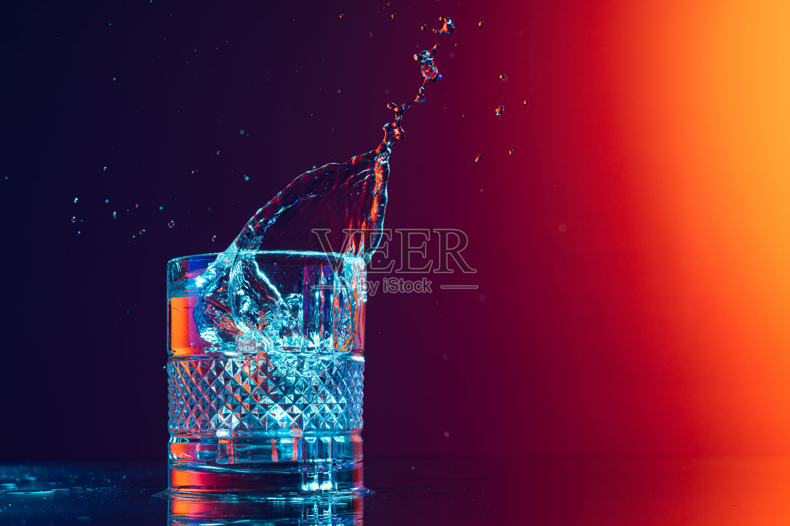 威士忌酒杯与冰块和水溅站在镜子表面上梯度黑橙背景在霓虹灯。艺术、美、饮料照片摄影图片
