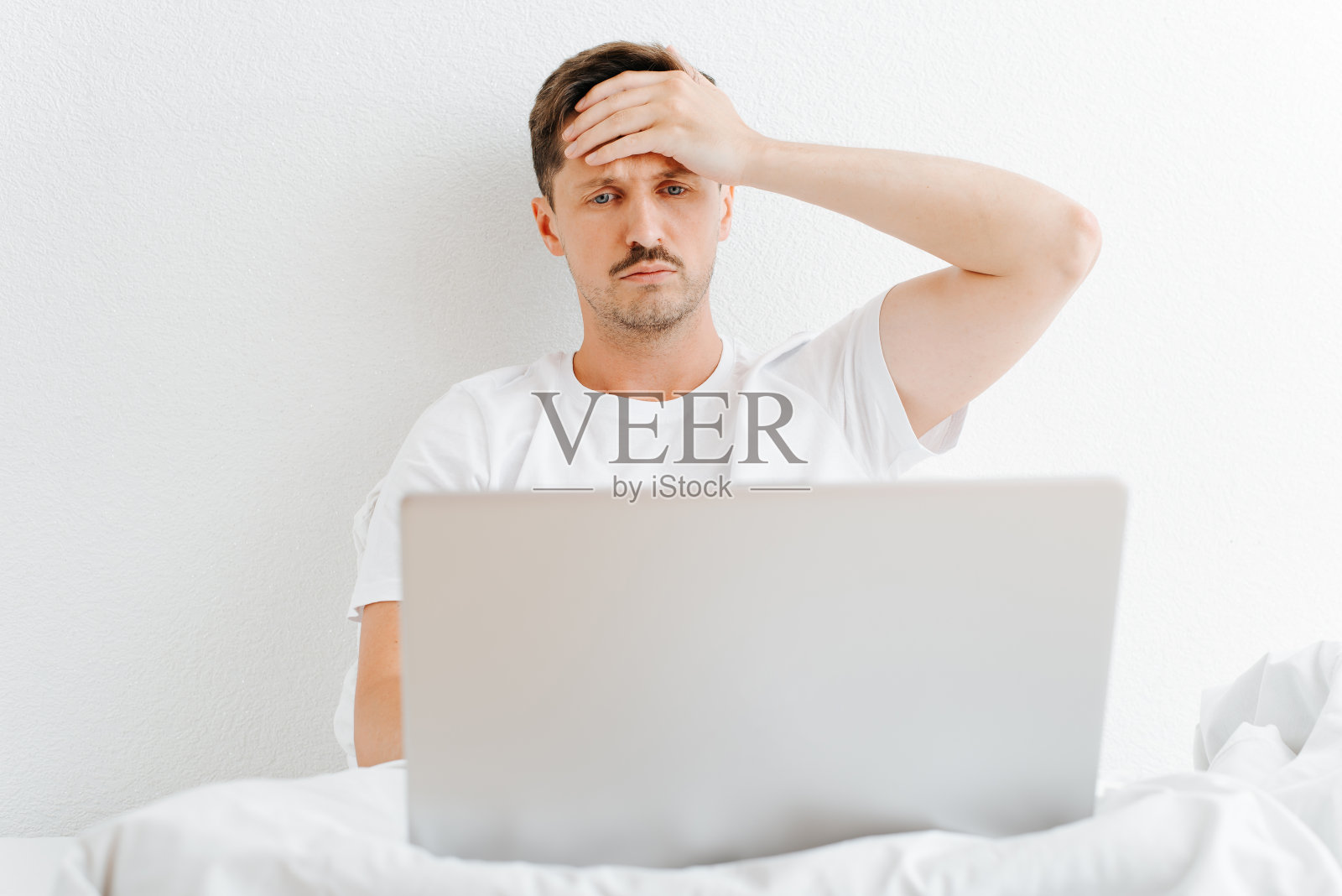 有头痛和疾病症状的疲倦男子坐在卧室的床上用笔记本电脑工作。悲伤的年轻人抱着头看着手机屏幕照片摄影图片