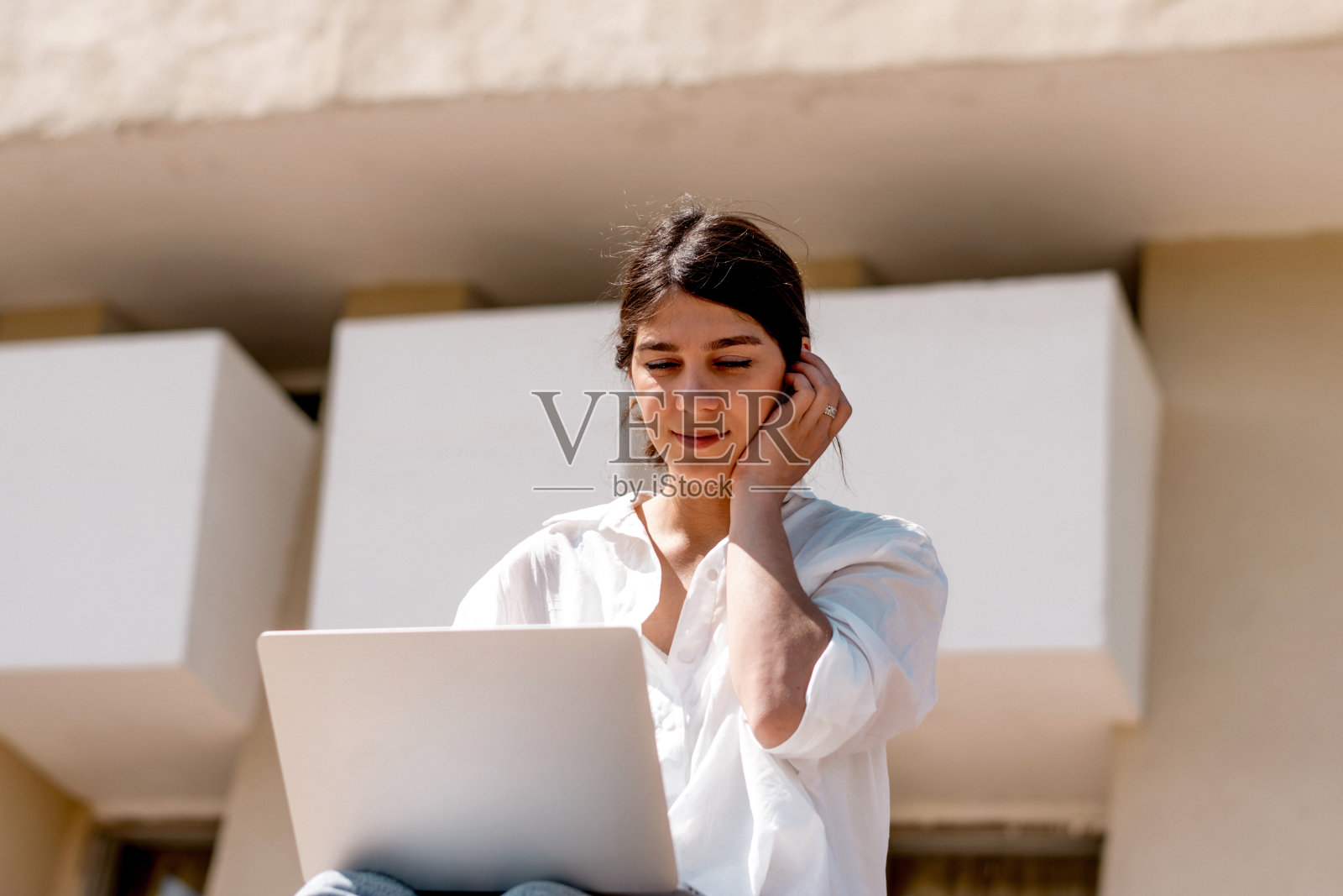 一个夏天,一位身穿白衬衫和牛仔裤的黑发年轻女子坐在城市街道的台阶上使用笔记本电脑。远程工作,自由职业,在线教育照片摄影图片