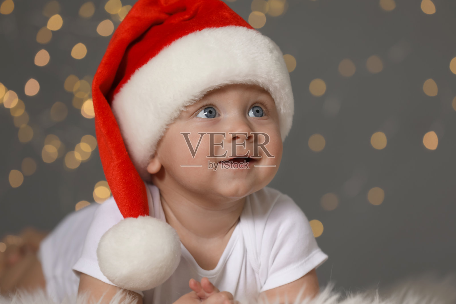 可爱的婴儿戴着圣诞帽,站在毛茸茸的地毯上,背对着迷离的灯光。圣诞节的庆祝活动照片摄影图片