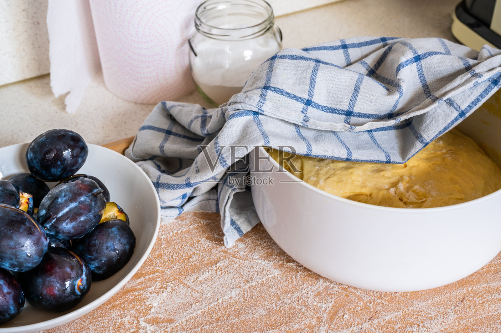 梅子放在盘子里,酵母面团放在碗里,用毛巾,玻璃杯里放糖,纸巾放在厨房木板上。照片摄影图片