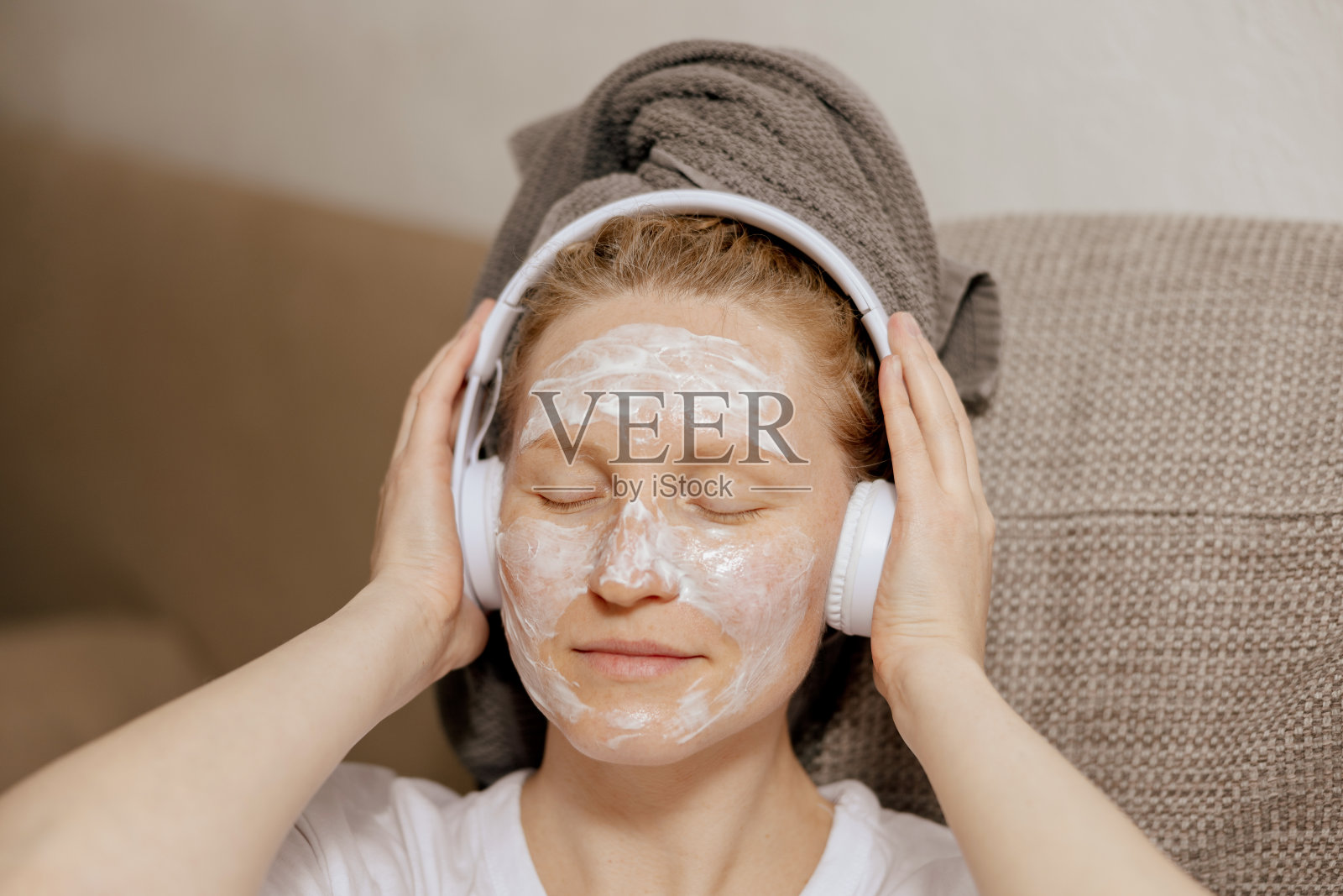 年轻女子穿着休闲装,头上戴着毛巾,脸上戴着美容面膜,坐在家里的沙发上,听着音乐休息。女性化妆品,皮肤护理。自己照顾自己,时间留给自己。放松。照片摄影图片