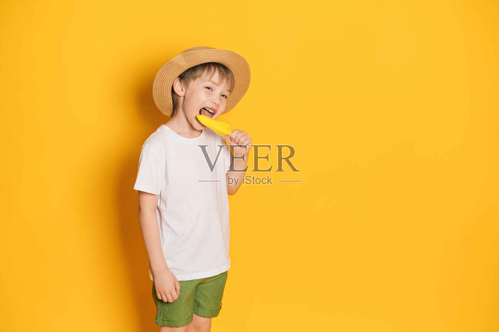 戴着帽子的男孩在黄色背景下舔着火箭形状的冰淇淋照片摄影图片