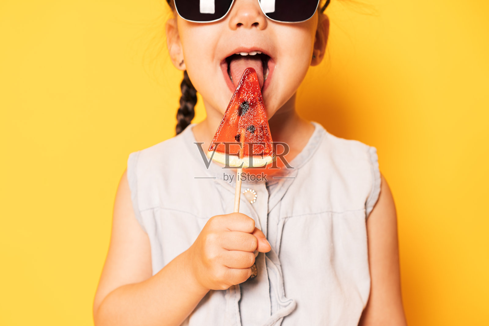 戴墨镜的女孩在黄色背景下舔着西瓜形状的棒棒糖照片摄影图片