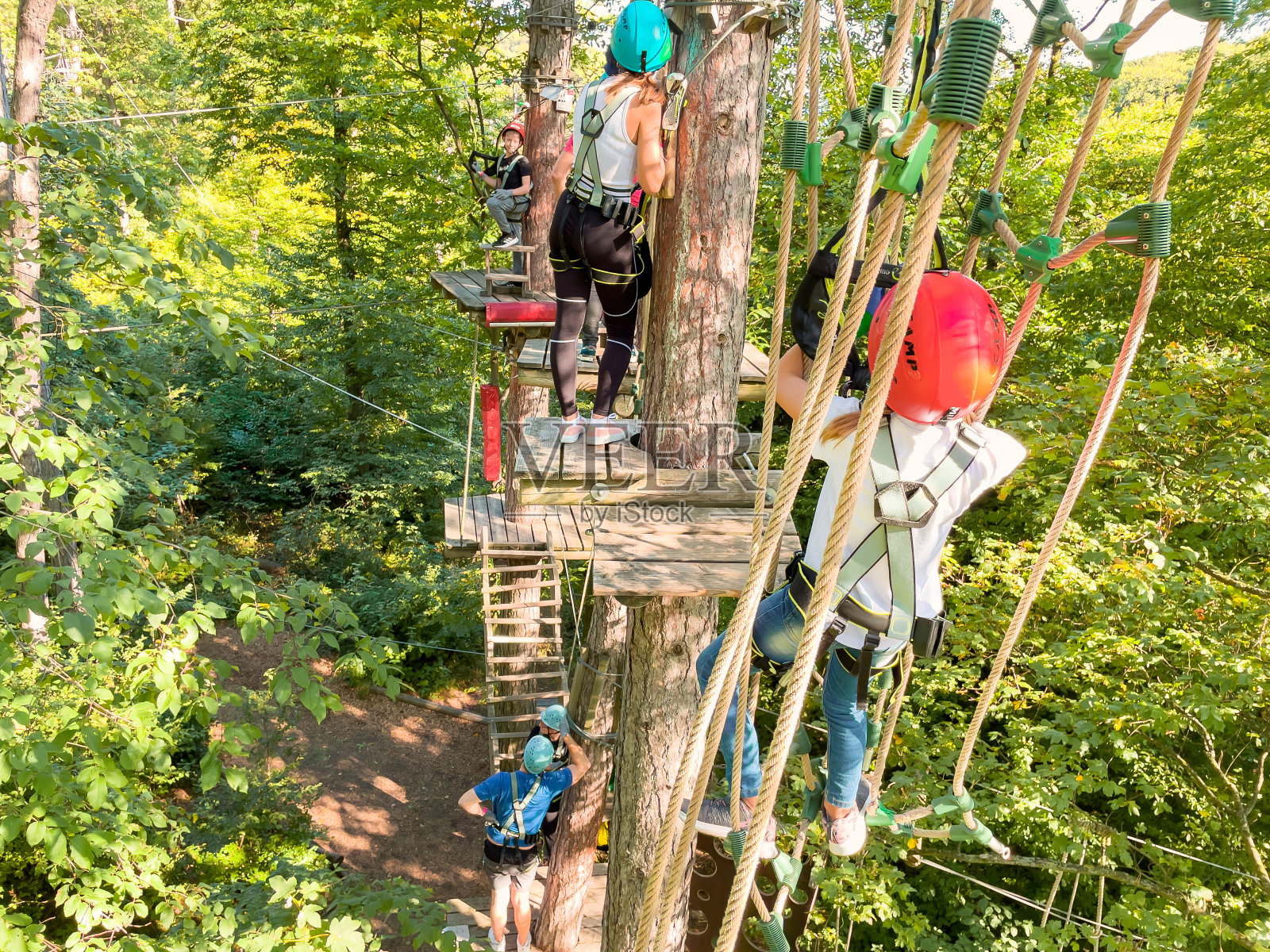 适合喜欢冒险的人的夏季娱乐和运动。高空攀登训练儿童在冒险轨道上,配备安全带和防护头盔。十几岁的女孩在夏季森林里的极限绳索公园的铰链小径上行走。照片摄影图片