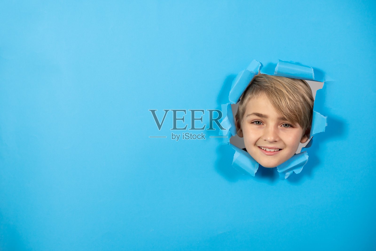 欢快的金发男孩的肖像与文本的复制空间。笑脸从蓝纸背景的一个洞里探出头来照片摄影图片