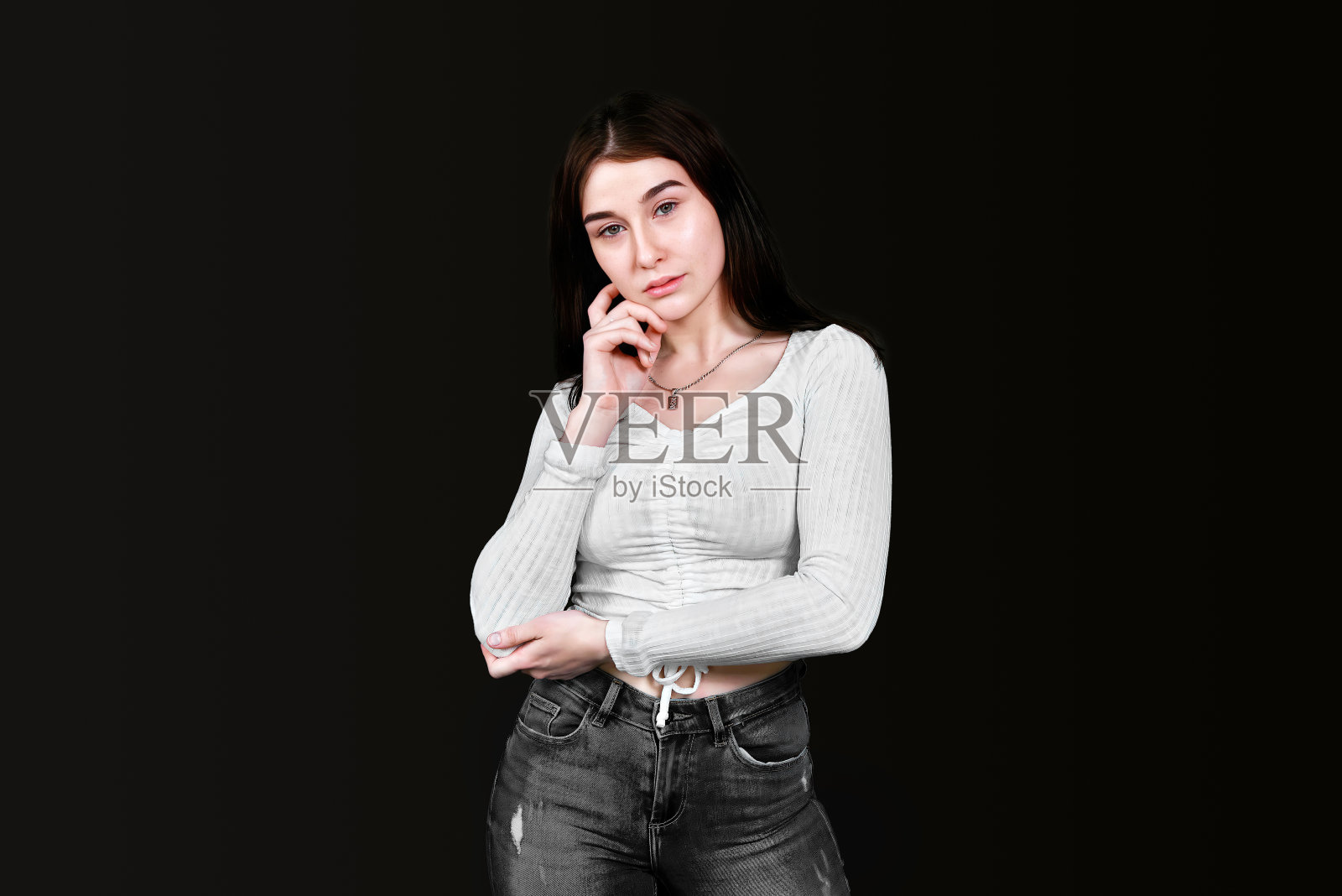 身材苗条的年轻女子身穿白色衬衫,胸部丰满,黑色牛仔裤,用手托着头,站在镜头前若有所思地看着镜头,背景是黑色。横幅,复制空间。照片摄影图片