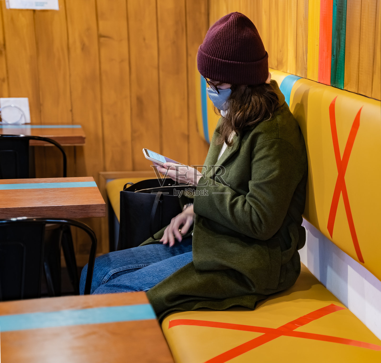 年轻的女孩,戴着防护口罩的女人来到明亮的咖啡馆。安全的社会距离。时尚的形象,外套,帽子,眼镜。标明禁区,餐厅客人的座位。冠状病毒大流行covid-19照片摄影图片