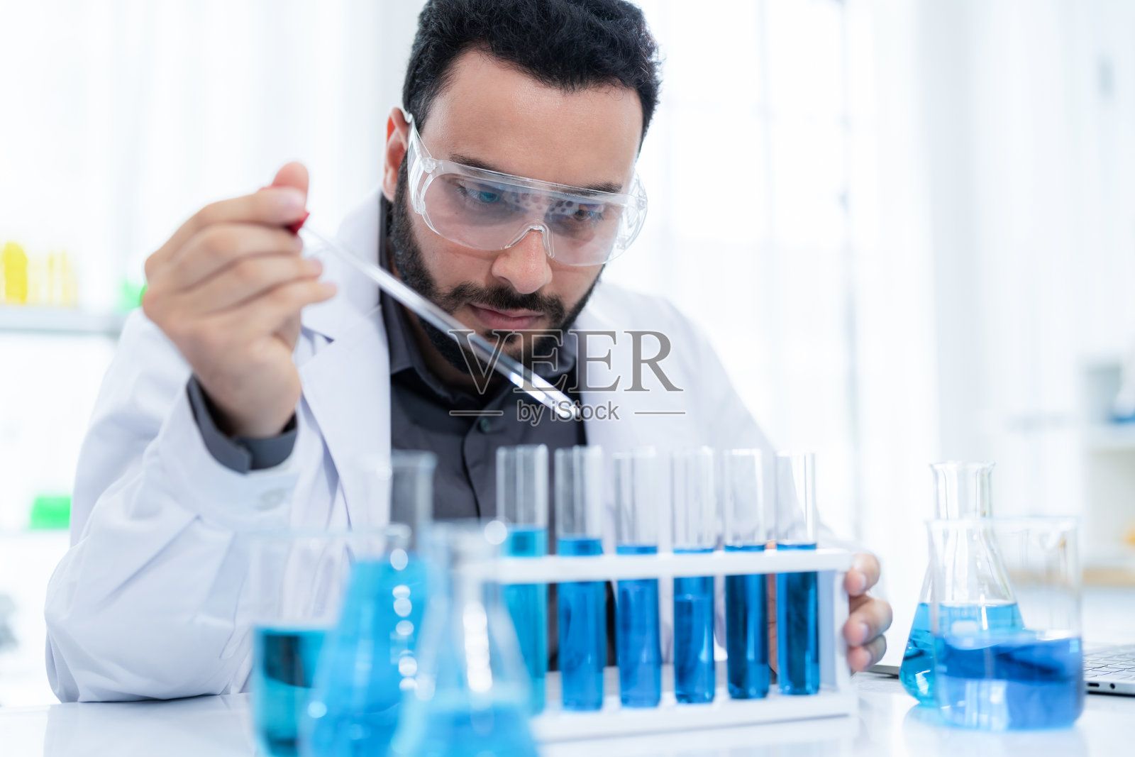 穿着白大褂、戴着安全眼镜的科学家将蓝色溶液滴入现代实验室的试管中。在笔记本电脑上记录,以便分析。研究发展,生物化学,医学,制药照片摄影图片