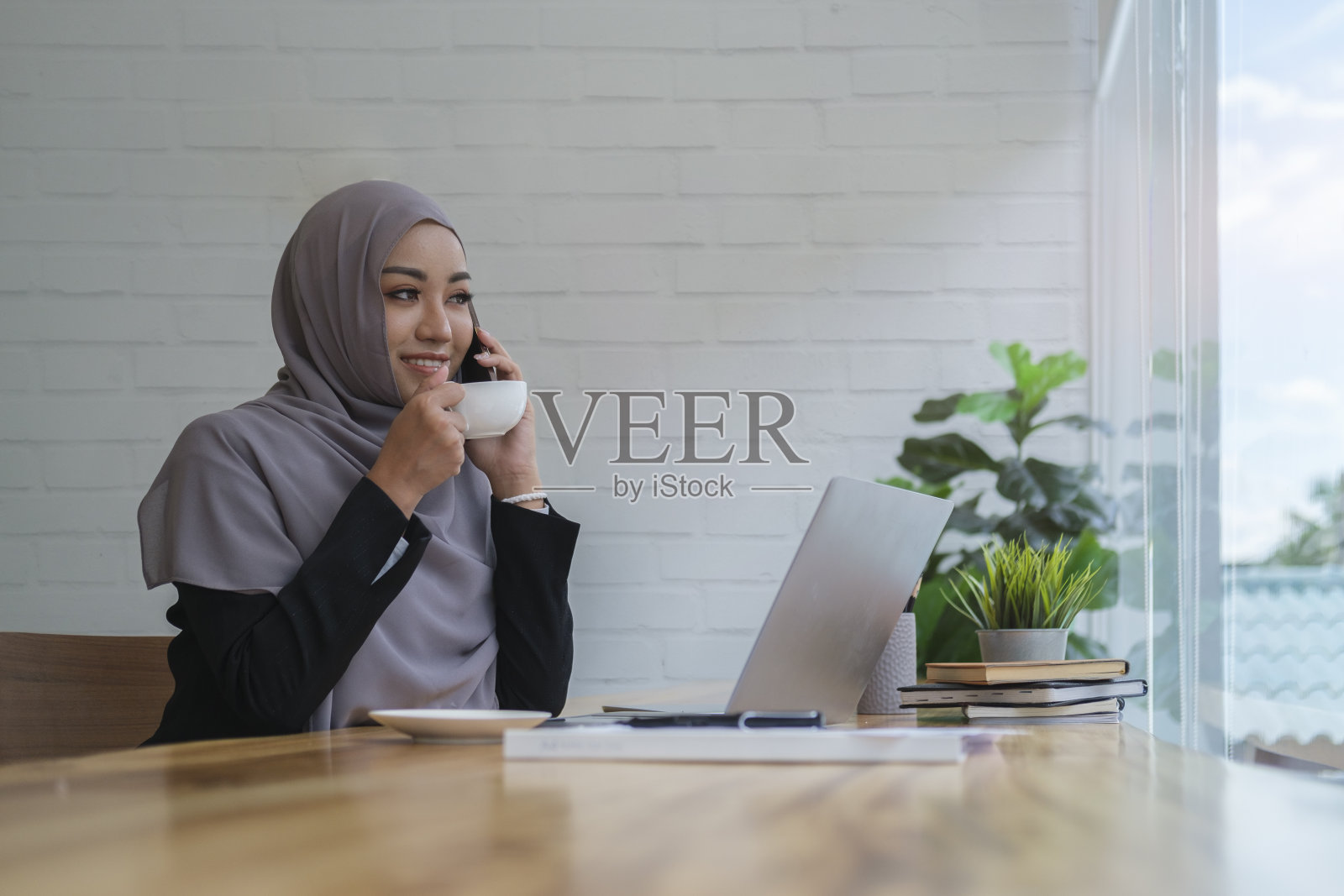 深思熟虑的穆斯林商业妇女喝着热咖啡坐在笔记本电脑前,看着窗外,想着工作计划。照片摄影图片