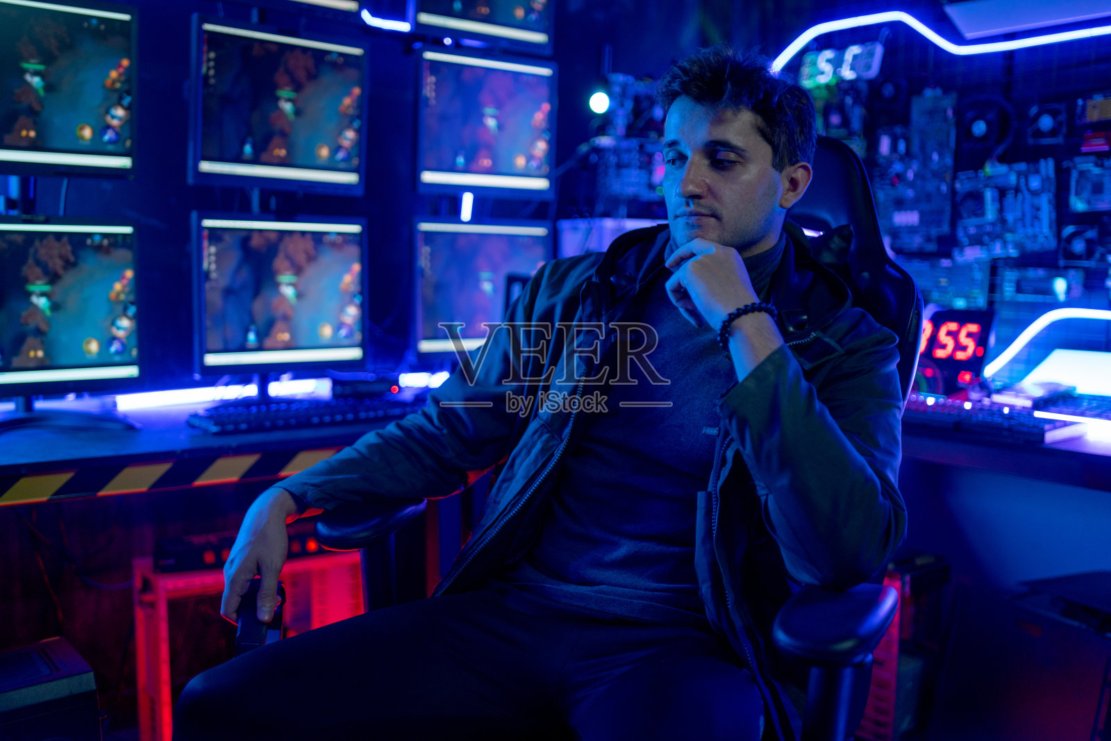 玩家男子坐着放松,把手指放在下巴上,看起来很舒服,在他的房间里多色的灯光播放游戏给粉丝俱乐部。照片摄影图片