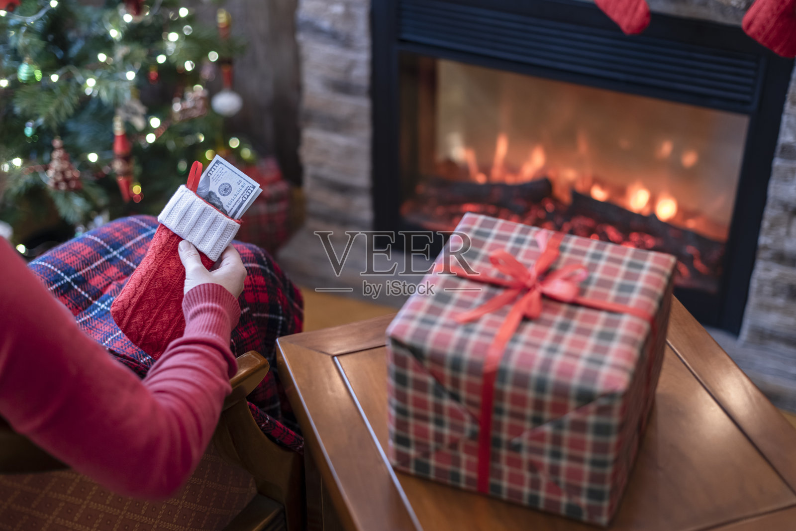女人把美元放在红色圣诞袜坐在圣诞树和壁炉旁边,包装礼盒。的概念。照片摄影图片