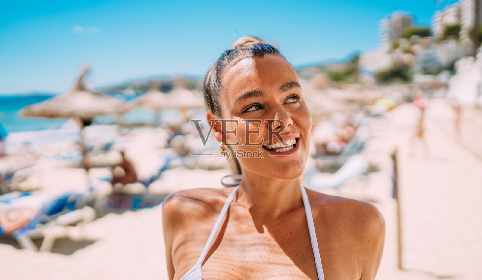 夏天沙滩上穿着比基尼的年轻欧洲女子。一个快乐的年轻女子对着大海微笑的画像。皮肤黝黑的金发女孩穿着白色泳装享受海滩。照片摄影图片