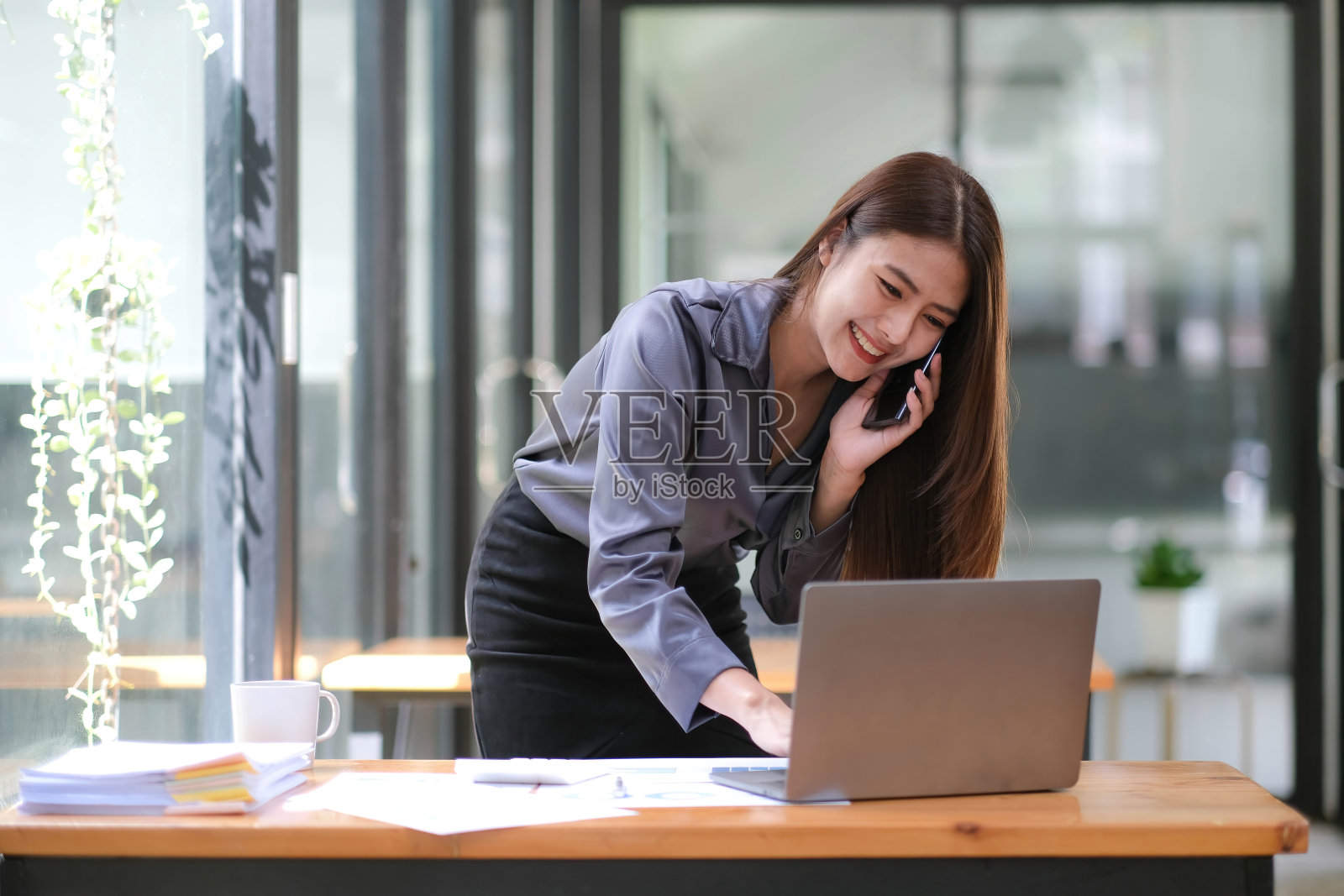 快乐的年轻亚洲女商人站在办公室使用智能手机和笔记本电脑。照片摄影图片