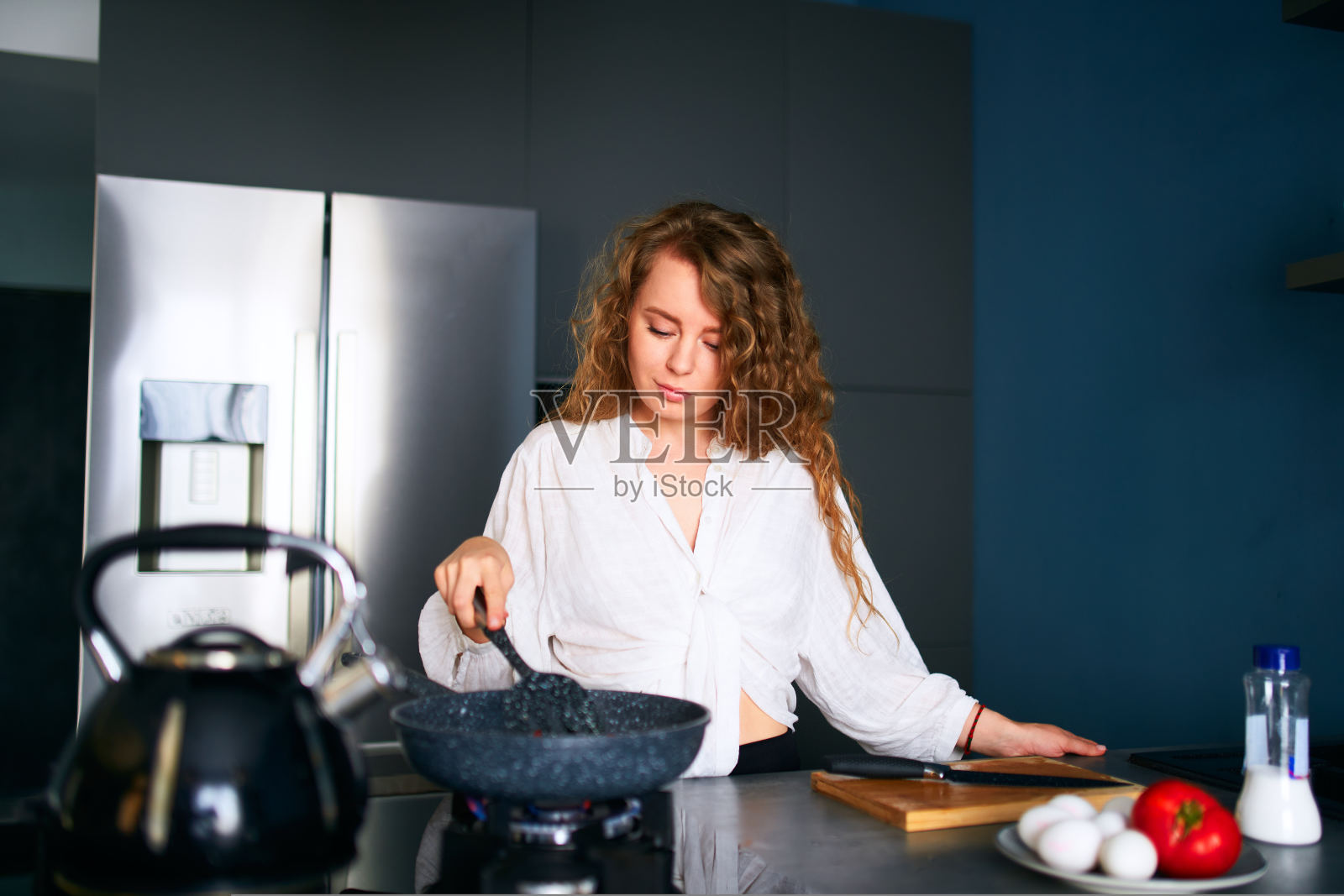 年轻微笑的卷发白人妇女在煎锅上做早餐。年轻的成年女性正在煤气炉上做煎蛋卷。女孩在一个现代简约的厨房岛上准备她的饭。煎照片摄影图片