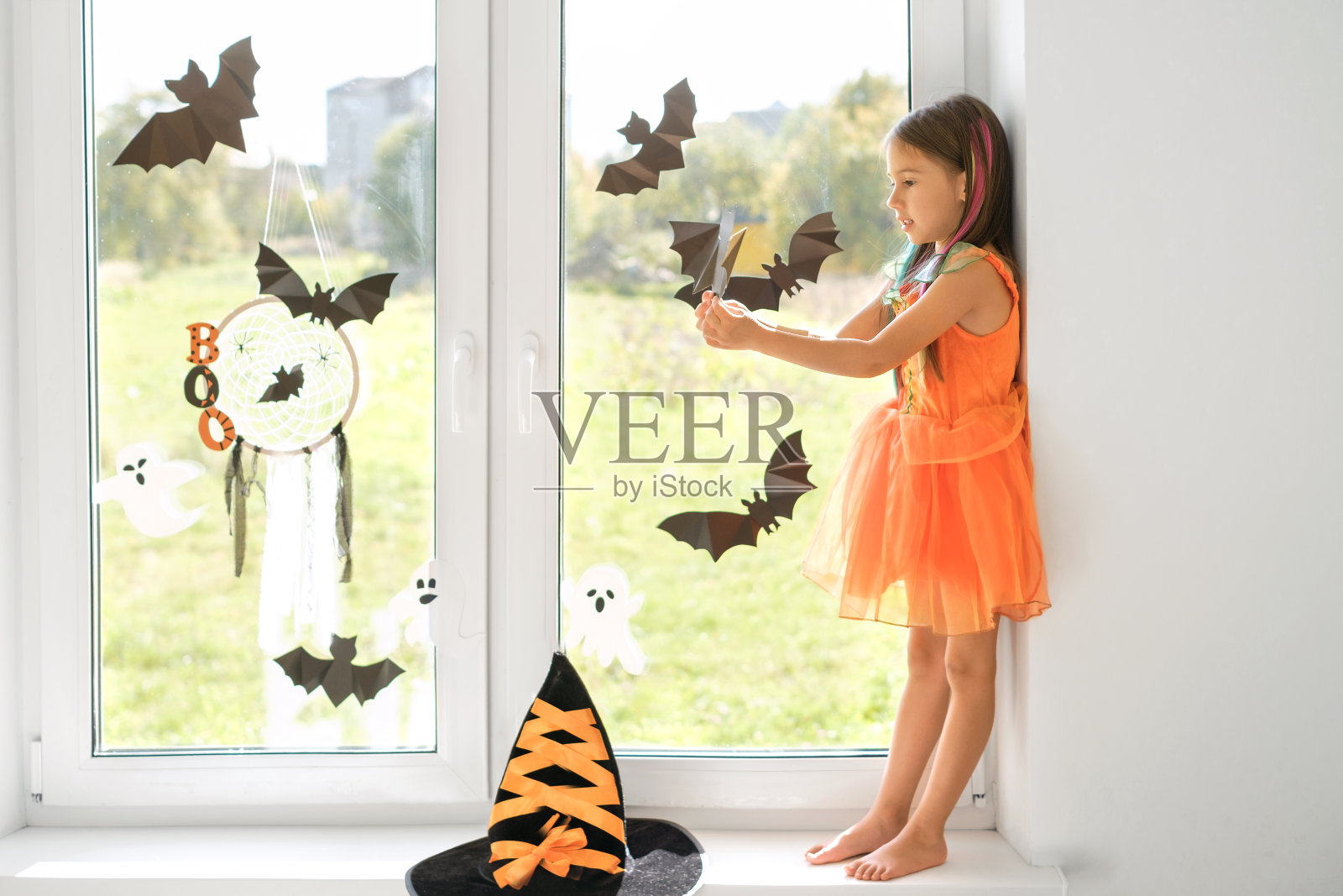 一个装扮成女巫的孩子站在窗台上,手里拿着一根纸球棒。万圣节庆典的准备工作。家居装饰照片摄影图片