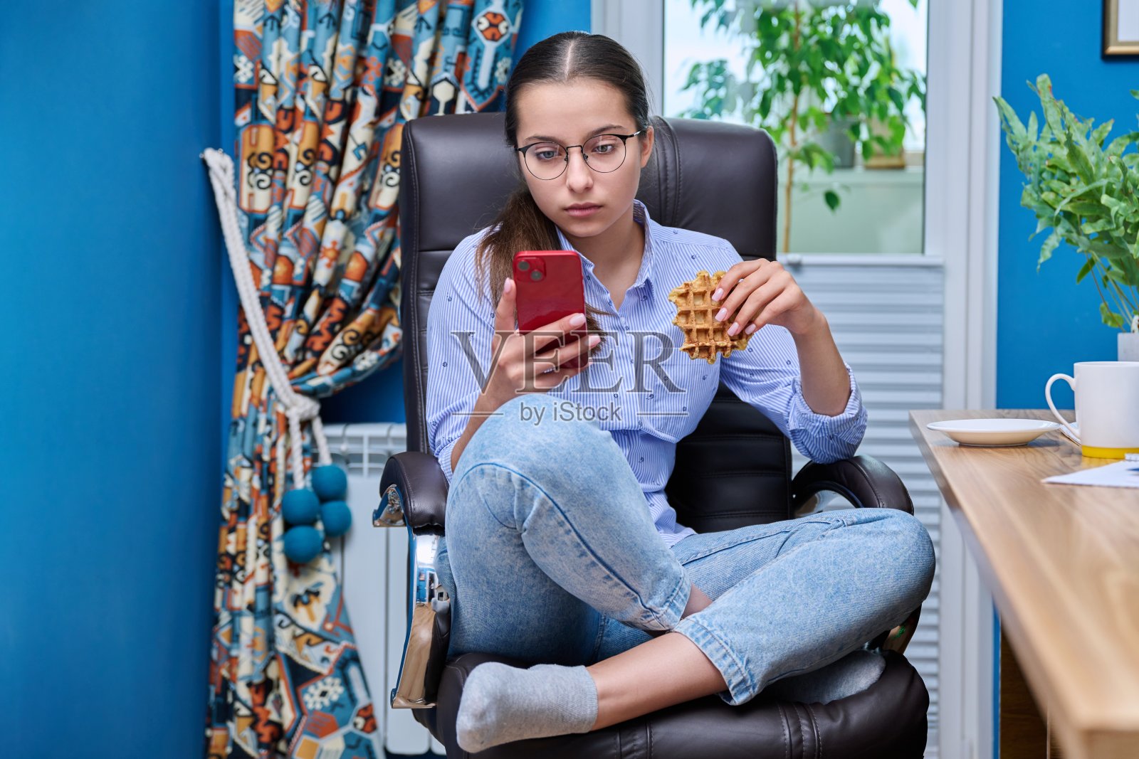 十几岁的女孩坐在电脑旁的扶手椅上,看着智能手机,吃着零食照片摄影图片