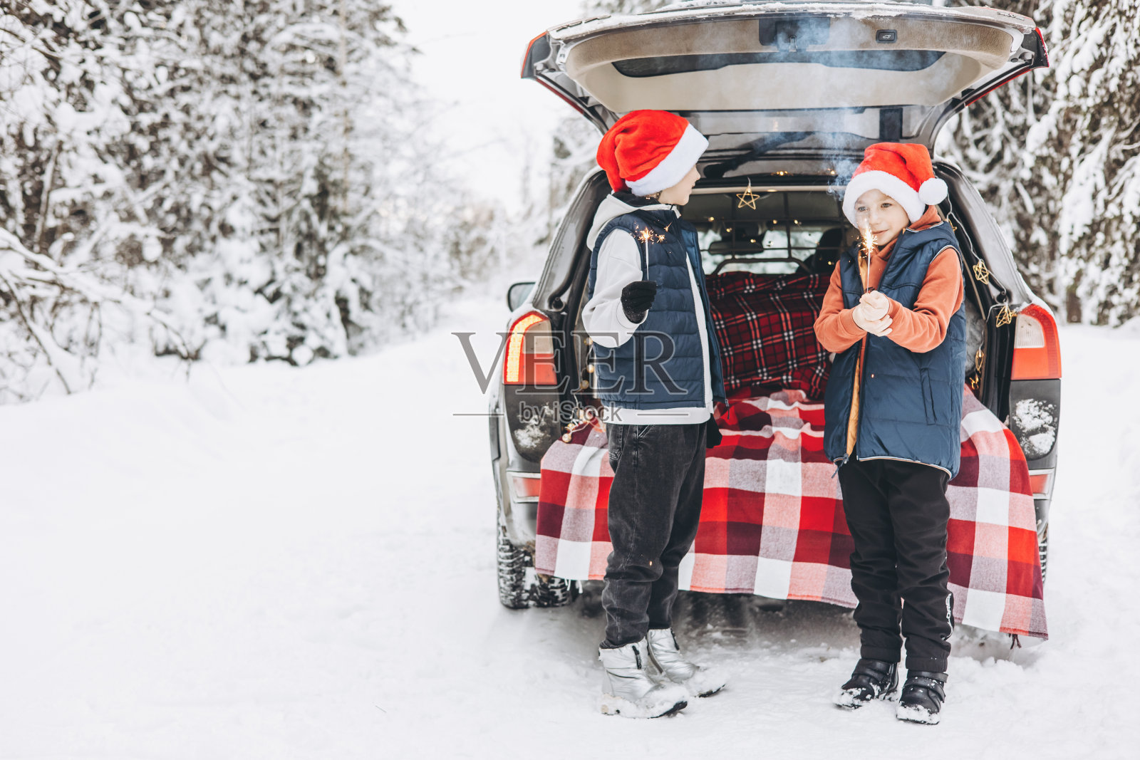 两个朋友,十几岁的男孩,戴着红色的圣诞帽,带着闪闪发光的孟加拉火,站在后备箱附近,为圣诞节和新年在下雪的冬天森林里装饰的汽车。自驾游和当地旅游。照片摄影图片