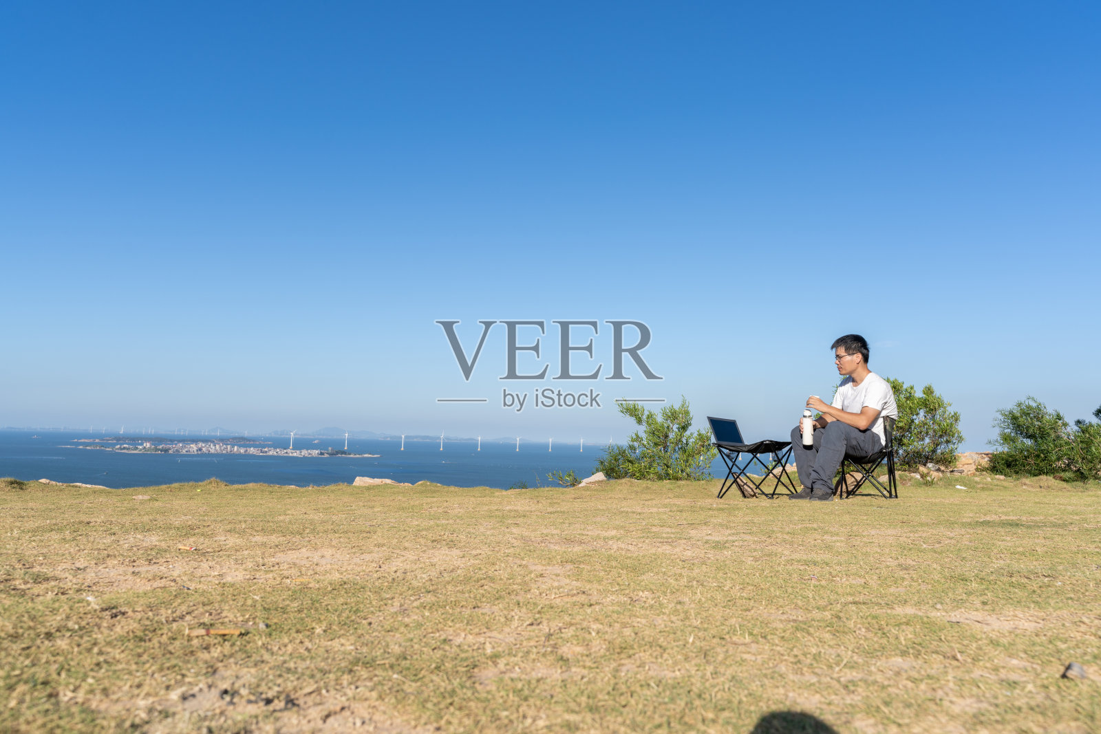 在野外的海边,一名亚洲男子坐在简单的桌椅上喝水照片摄影图片