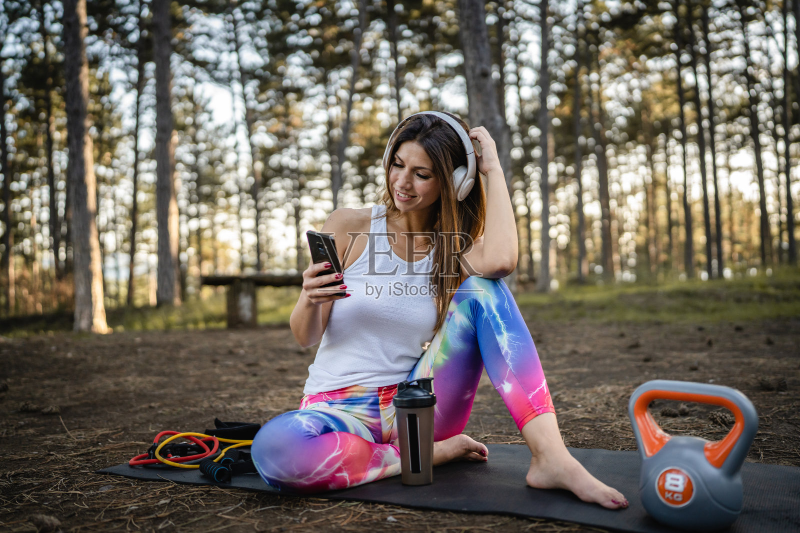 一名女性,成年白人女性,在夏天的森林或公园里训练时使用手机刹车,用壶铃设备听耳机和手机应用程序的音乐照片摄影图片