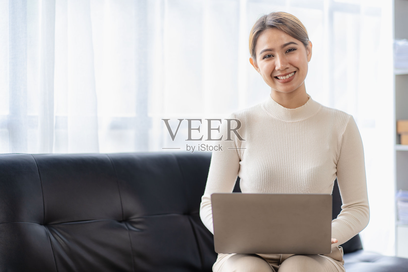 亚洲妇女坐在沙发上,带着笔记本电脑微笑,开心地上网和网上购物。照片摄影图片