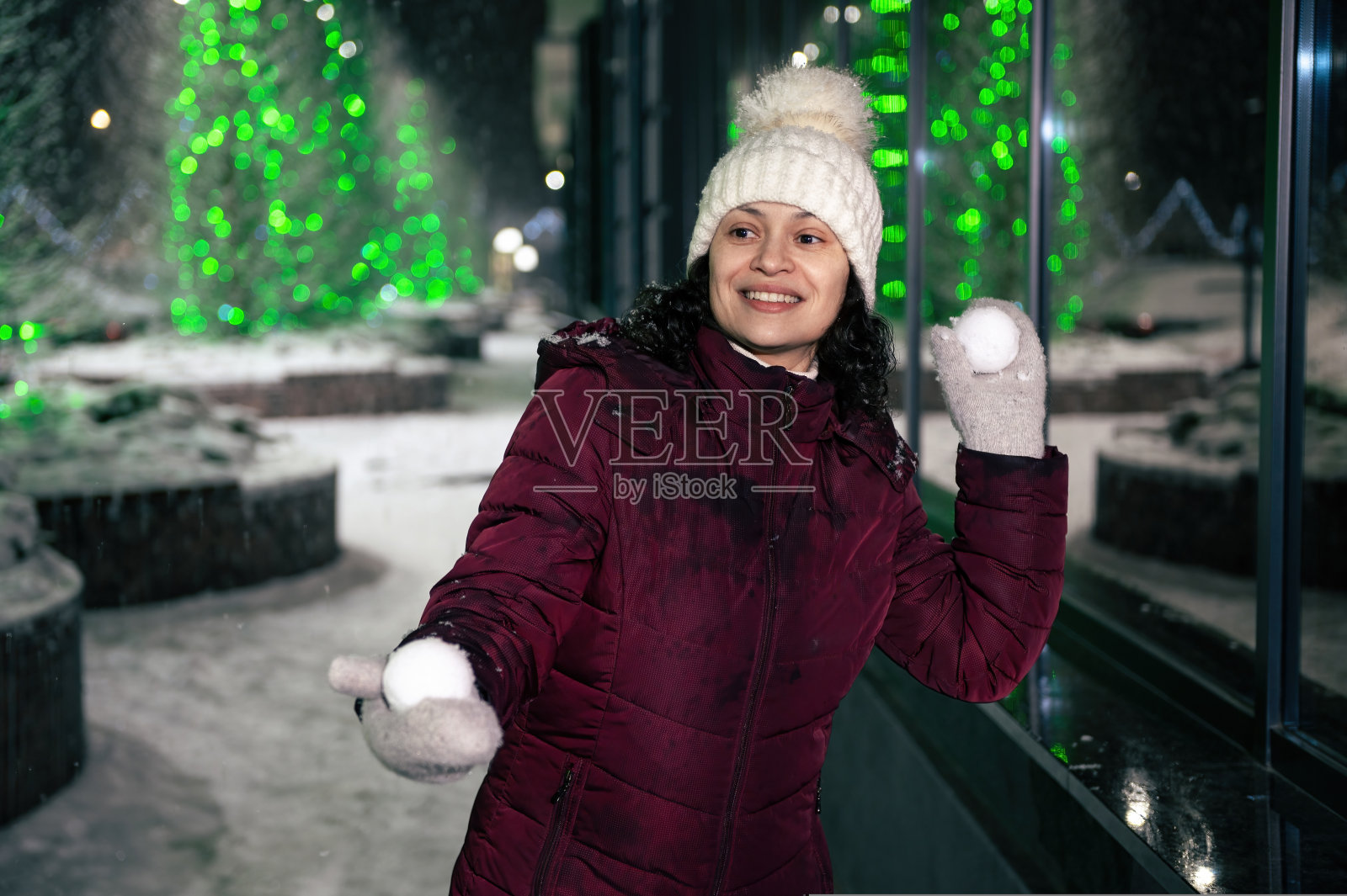 快乐的年轻女子穿着暖和的衣服,戴着白色的羊毛帽子和手套,在一个下雪的冬夜打雪仗照片摄影图片
