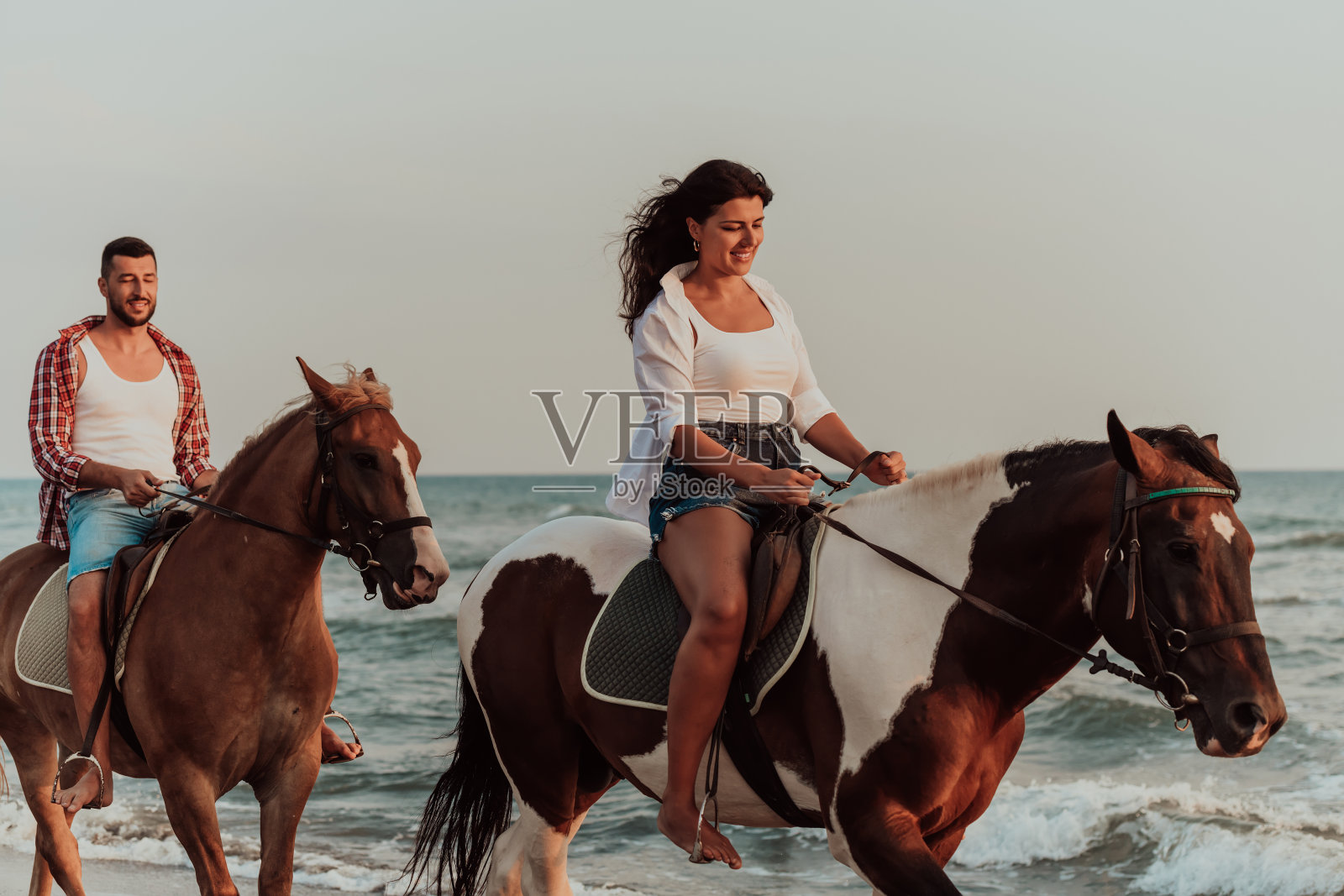 夕阳西下,一对相爱的情侣穿着夏装在沙滩上骑马。背景是大海和日落。有选择性的重点照片摄影图片