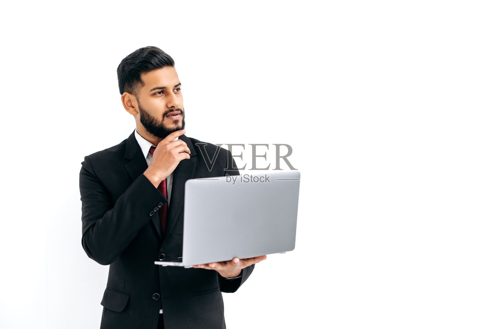 困惑的深思熟虑的优雅有影响力的阿拉伯或印度男人穿着正式的西装,站在孤立的白色背景上,拿着笔记本电脑,沉思地看向另一边,思考,梦想。演示文稿的复制空间照片摄影图片