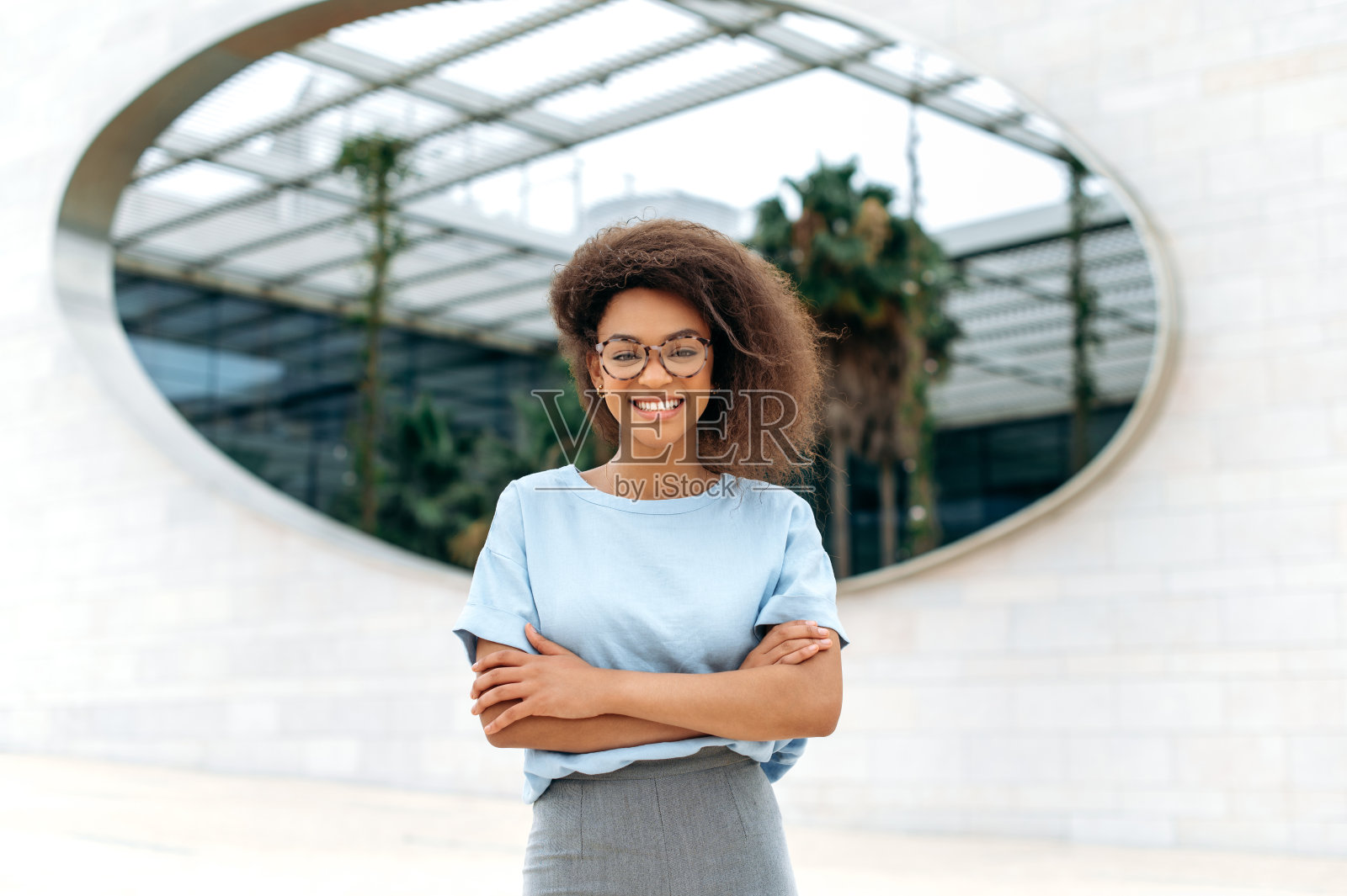 画像自信成功的非裔美国卷发商业女性,穿着优雅时尚的衣服,戴着眼镜,站在商业中心附近的户外,双臂交叉,看着相机,微笑照片摄影图片