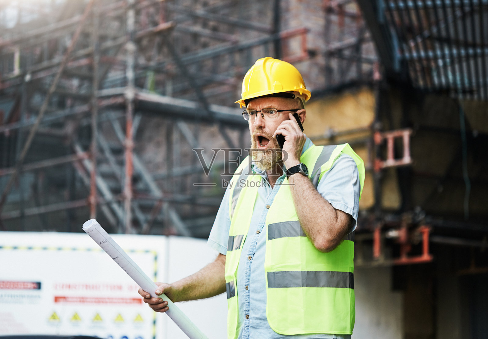 建筑工人拿着建筑图纸站在建筑工地上,从手机里听到坏消息照片摄影图片