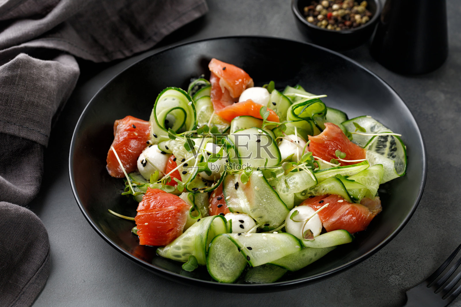 三文鱼沙拉配黄瓜片,马苏里拉奶酪,青菜放在黑色的盘子和灰色的背景上。前视图。照片摄影图片