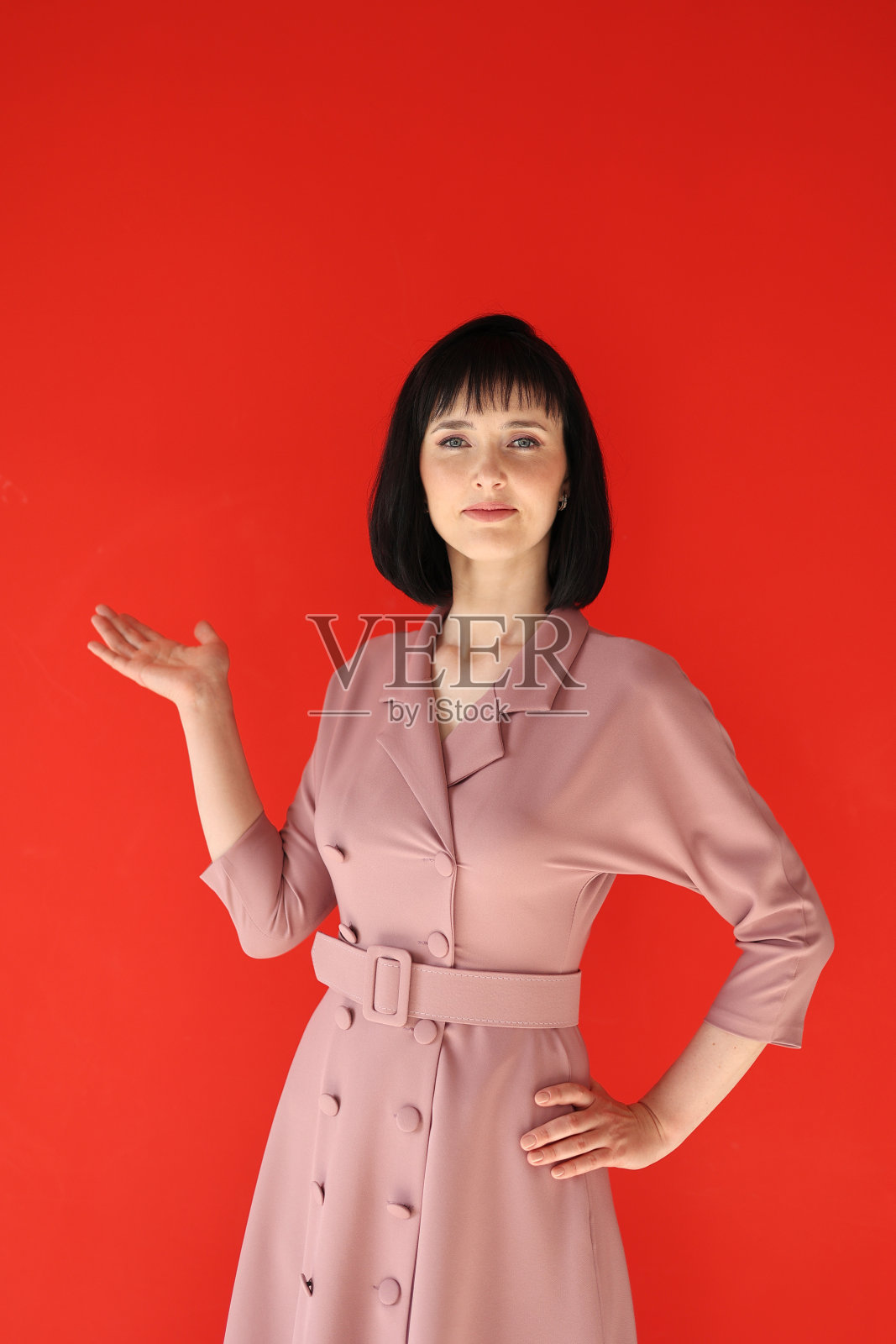 美丽的褐发年轻的商务女性穿着粉红色的套装看着相机指着手在复制空间孤立在红色的工作室背景,微笑美丽自信的女士专业展示一旁照片摄影图片