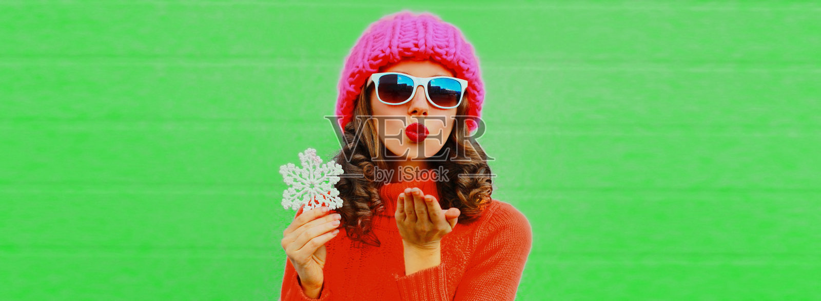 冬天的肖像美丽的年轻女子与雪花吹她的嘴唇针织毛衣,粉红色的帽子在绿色的背景照片摄影图片