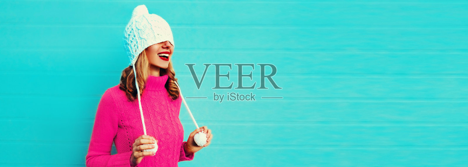 冬天的肖像,快乐的微笑年轻女子穿着粉红色的针织毛衣,白色的帽子在蓝色的背景上玩得很开心照片摄影图片
