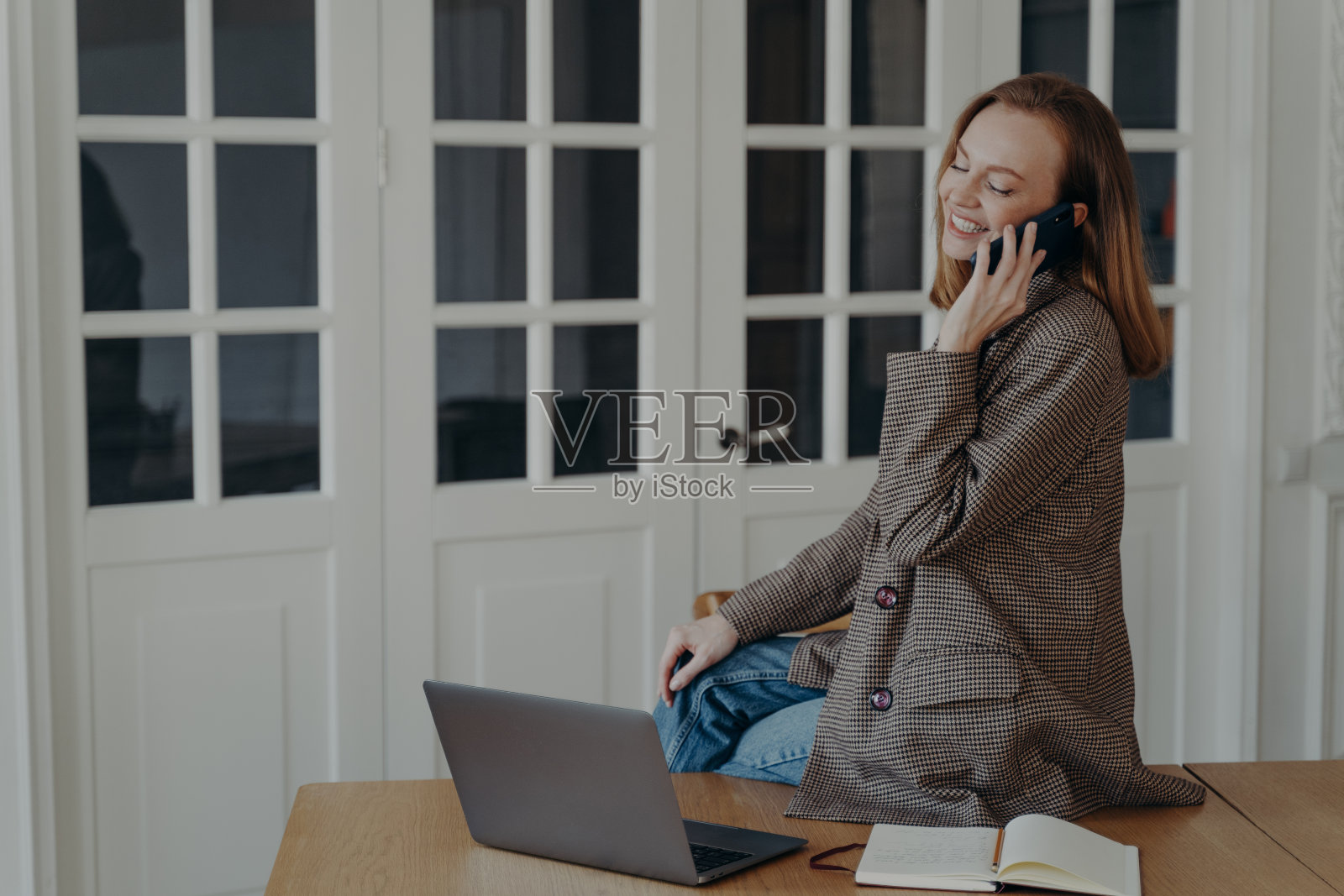 面带微笑的自由职业女经理接听手机电话,坐在桌子上用笔记本电脑打电话照片摄影图片
