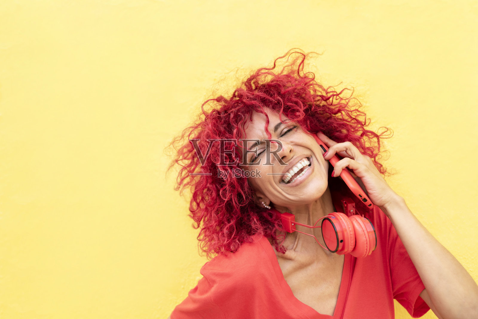 一个快乐的拉丁妇女的肖像,红头发的爆炸头在她的手机上说话和大笑,红色耳机在她的脖子上,黄色背景照片摄影图片